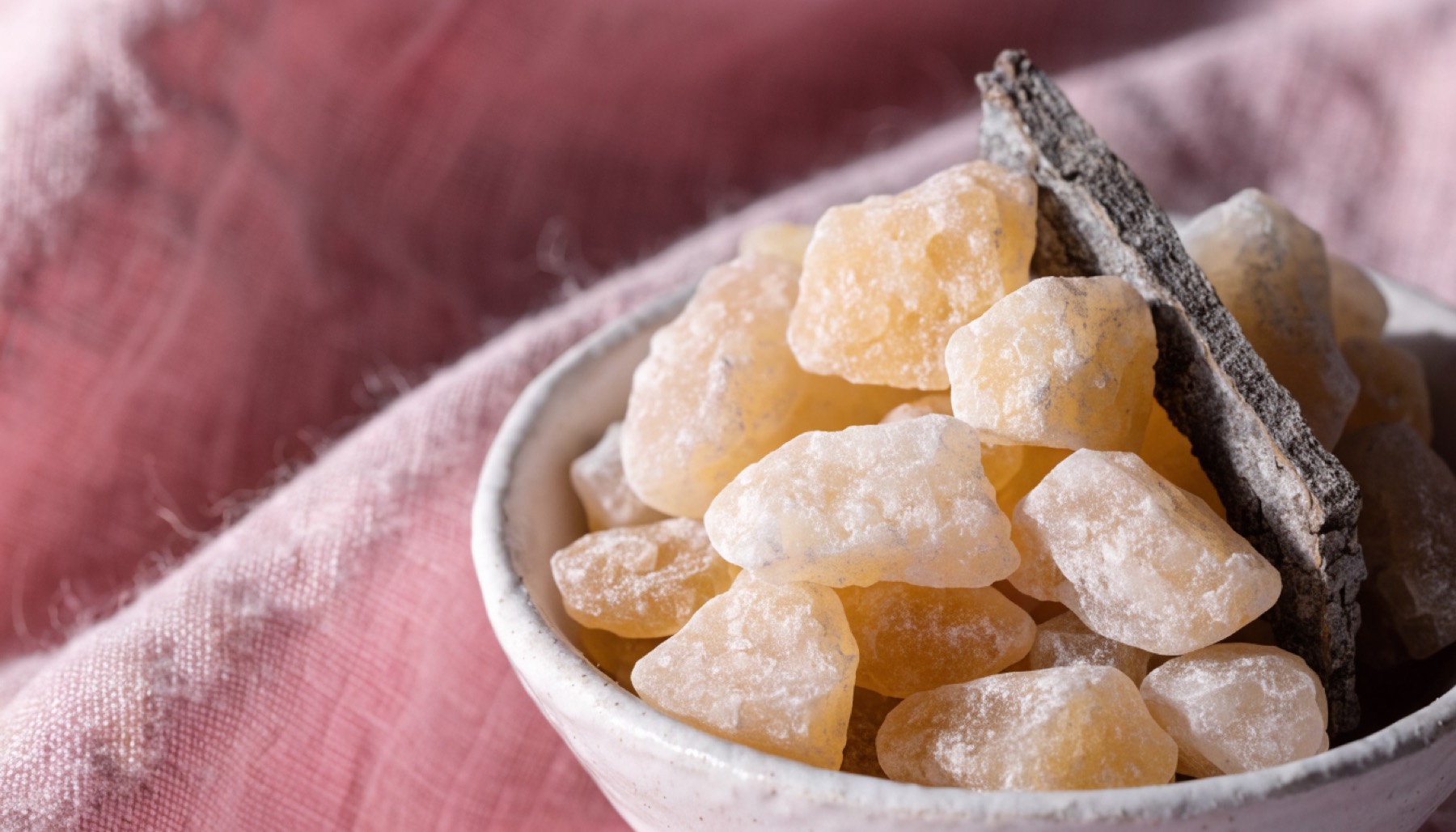 Lágrimas de resina dorada y ámbar claro amontonadas en un pequeño cuenco de cerámica blanca sobre un paño de lino rosa